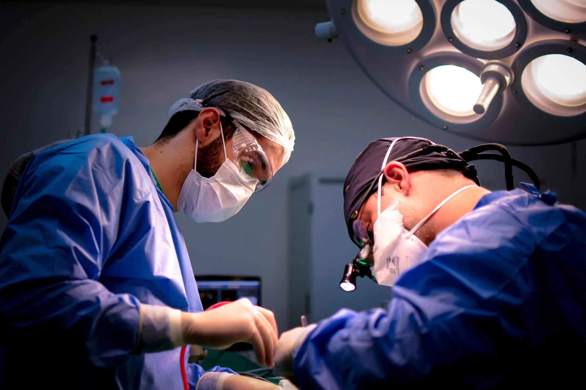 Dois cirurgiões concentrados durante uma cirurgia, com foco de luz frontal, óculos de proteção e lupas cirúrgicas