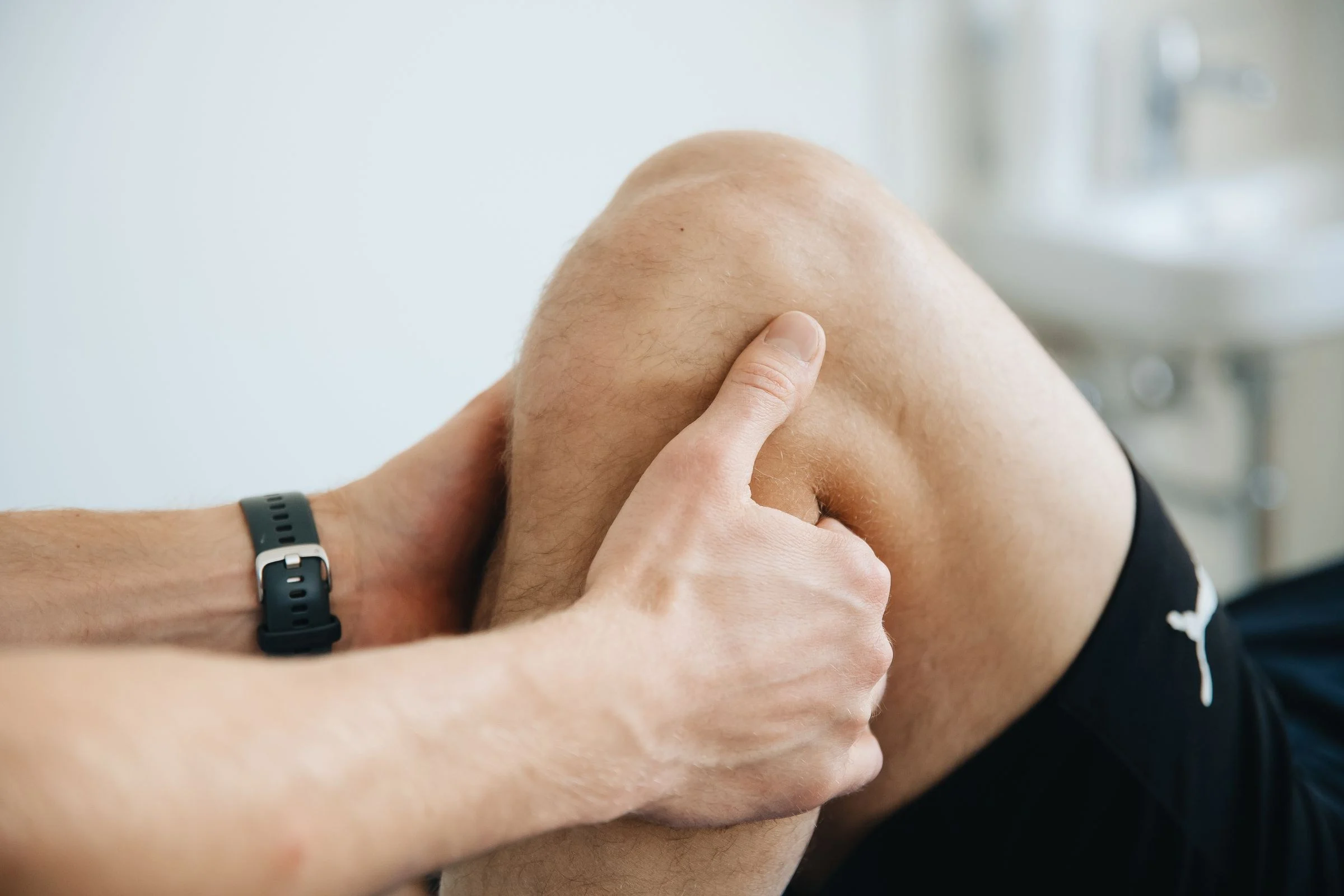Mãos de um profissional palpando o joelho flexionado de um paciente durante o exame clínico