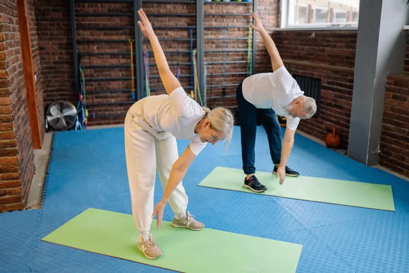 Casal de idosos fazendo alongamento de tronco sobre colchonetes em sala de exercícios
