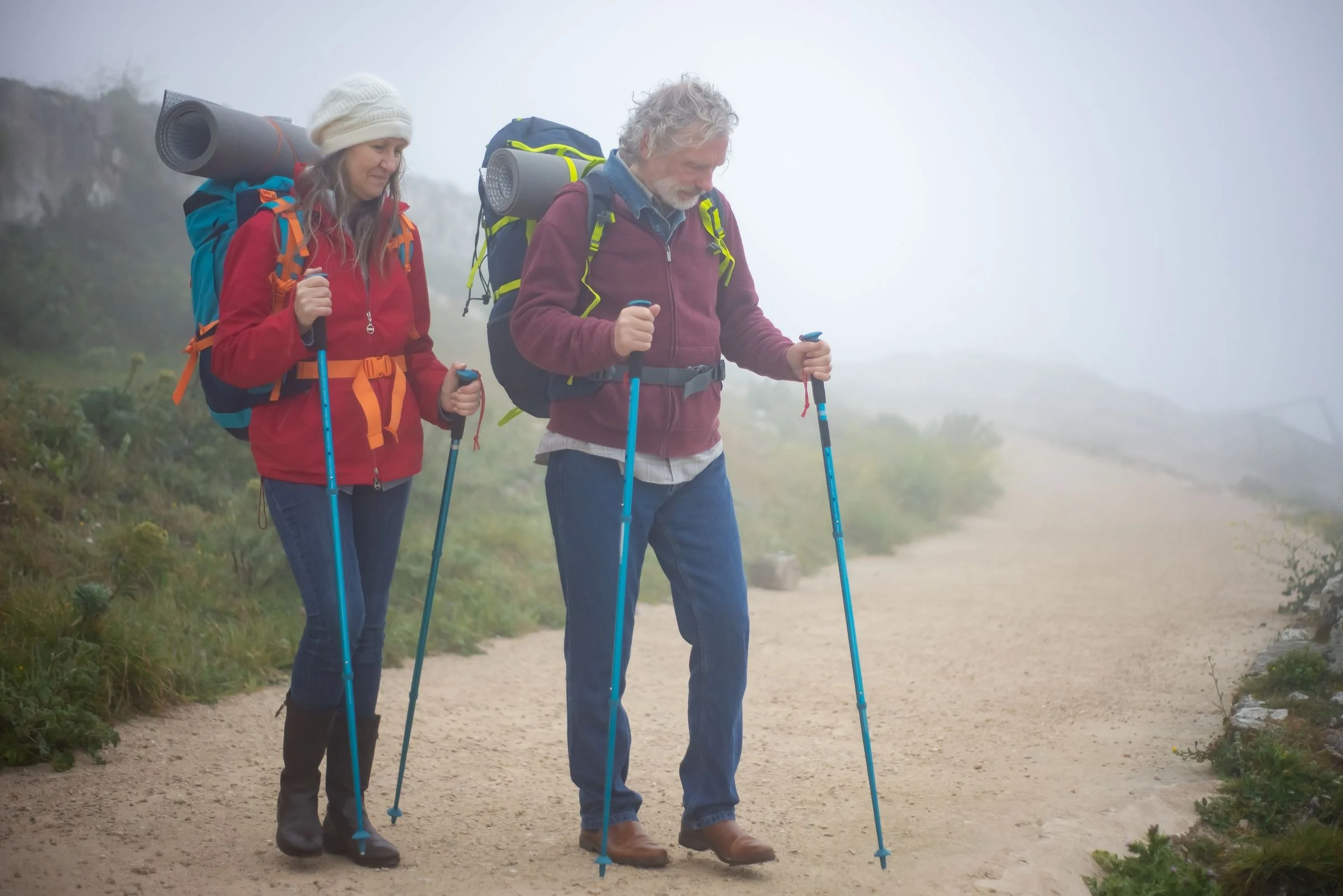 Casal de meia-idade caminhando numa trilha de terra, com mochilas e bastões de caminhada